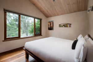 A double bed in a cream rendered bedroom with a large wooden window overlocking the bush